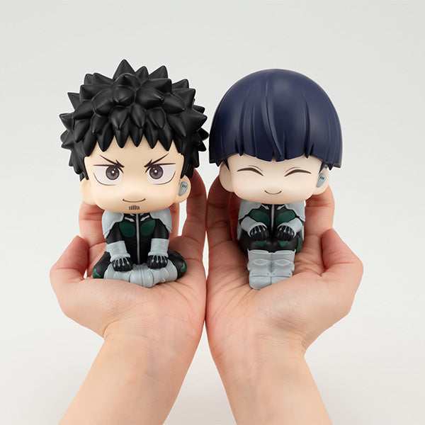 Two collectible figures held in hands, one with spiky black hair and the other with short dark hair, both wearing green and gray outfits and sitting in a pose looking upwards.